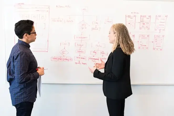 Two people discussing in front of a whiteboard filled with diagrams and charts